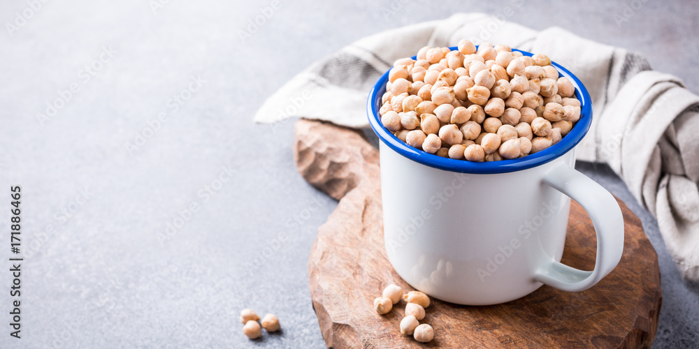 Raw uncooked organic chickpeas in white enamel mug on rustic wooden board on light gray stone background. Healthy vegan vegetarian food concept. Banner. Raw uncooked organic chickpeas in white enamel mug on rustic wooden board on light gray stone background. Healthy vegan vegetarian food concept. Banner.