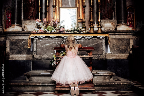 a LIttle blonde girl in a pink dress praying near the altar in the temple. View from behind.the girl kneels in church