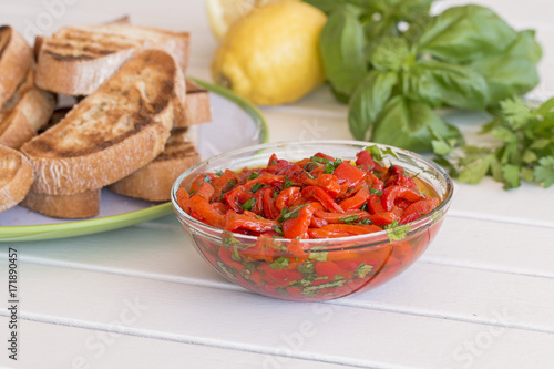 A glass bowl with red sweet pepper, a plate with some bruschetta. Background: lemon, parsley and basil.
