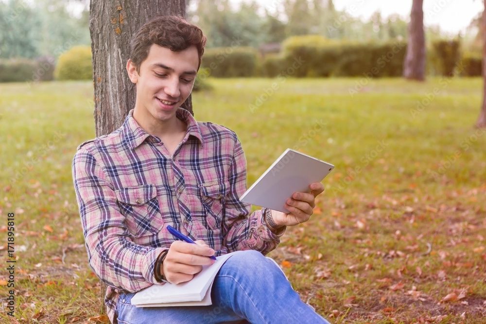young men student sitting in the park next to the tree with the tablet in his hands young men student sitting in the park next to the tree with the tablet in his hands