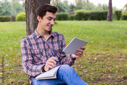 young men student sitting in the park next to the tree with the tablet in his hands