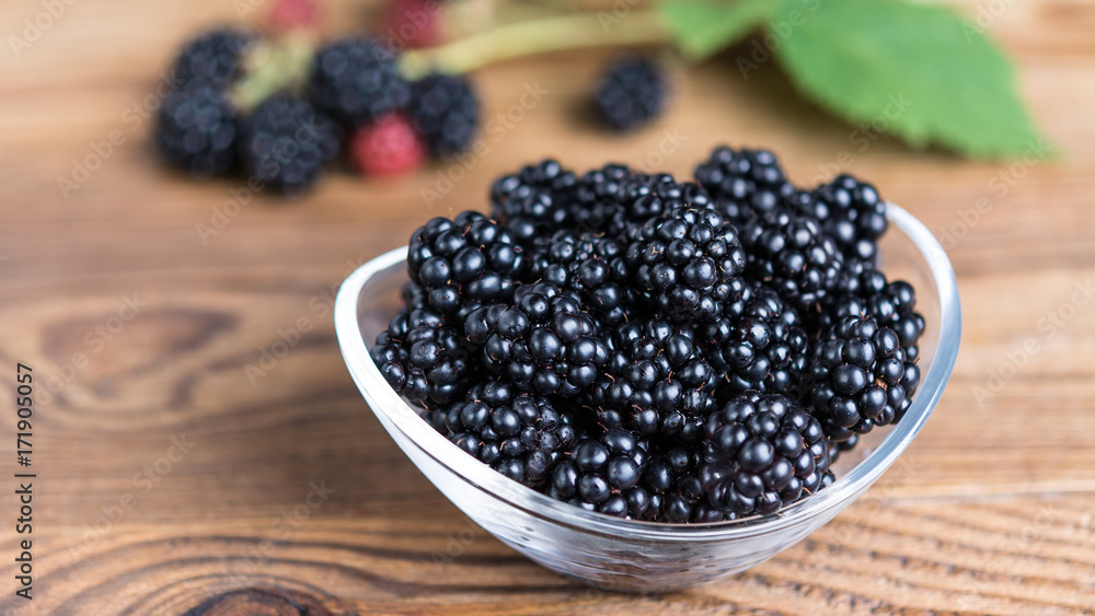 Sweet juicy blackberries in a glass bowl. Fresh forest fruits on wooden table with blurred bramble branch in the background. HD ratio 16x9. Sweet juicy blackberries in a glass bowl. Fresh forest fruits on wooden table with blurred bramble branch in the background. HD ratio 16x9.