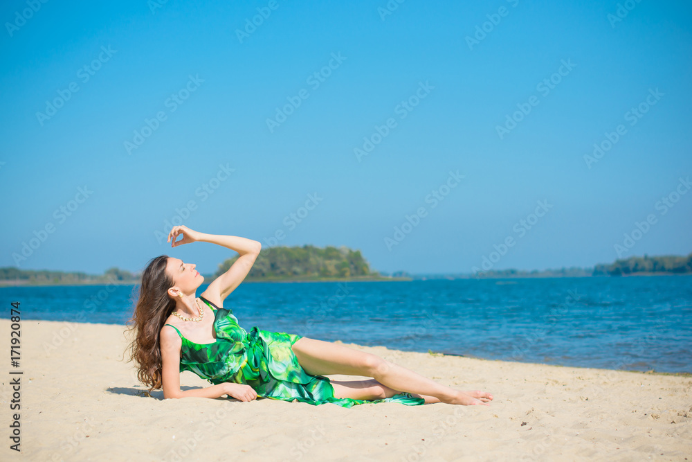 Woman wearing in green dress rest on a beach at warm day.  Mature thin lady Outdoors  on beach sand near water Landscape. Attractive Middle Aged Woman rest on nature