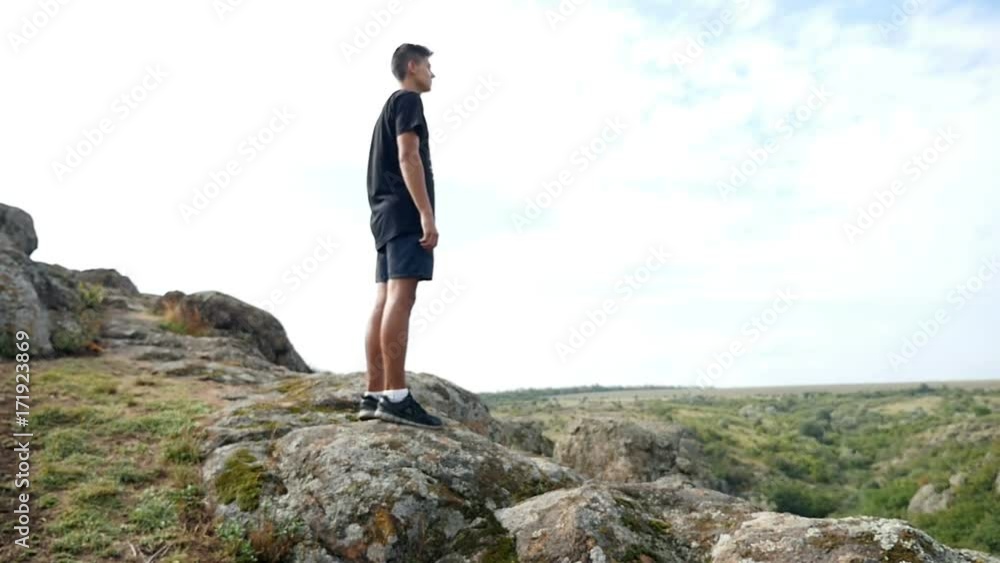 Impressive view of young man, dressed in black shirts and a t-shirt, while standing on a top of a cliff looking on picturesque hills in summer in slow motion. The landscape is really great Impressive view of young man, dressed in black shirts and a t-shirt, while standing on a top of a cliff looking on picturesque hills in summer in slow motion. The landscape is really great