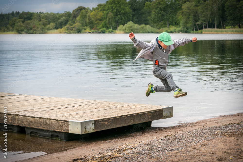 Naklejka premium Young playful caucasian boy running in mid-air making a jump from a jetty to the beach.