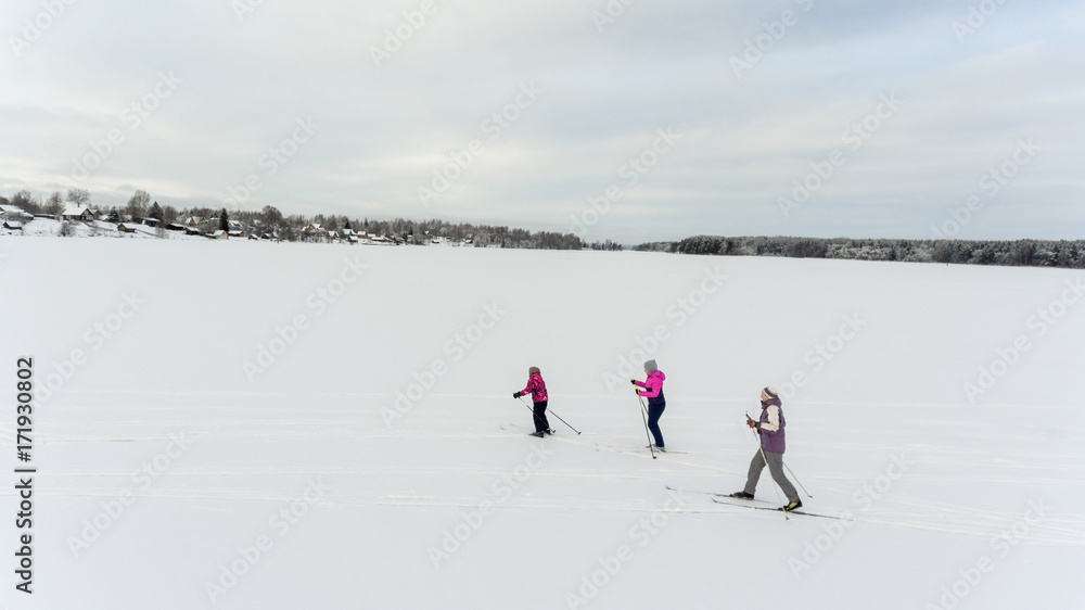 Three people skiing in frozen snowy lake to the village on shore, winter in Russia