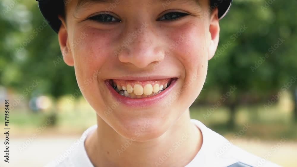 A happy eleven-year-old boy in a cap smiles while standing in a picturesque park with a lot of green trees and beautiful lawn on a sunny day in summer in slow motion A happy eleven-year-old boy in a cap smiles while standing in a picturesque park with a lot of green trees and beautiful lawn on a sunny day in summer in slow motion