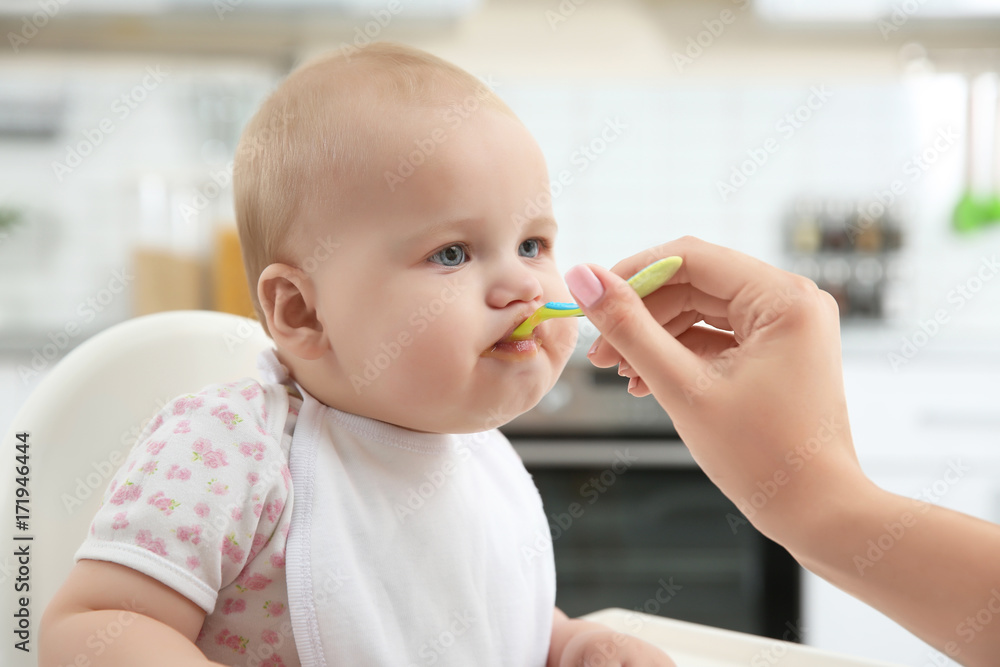 Mother feeding baby with spoon in kitchen Mother feeding baby with spoon in kitchen