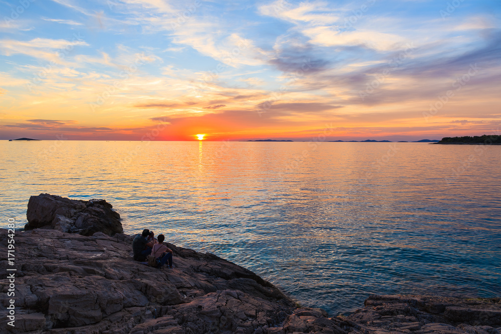 Unidentified couple of people sitting on rocks and watching sunset in Primosten town, Dalmatia, Croatia