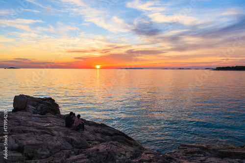 Unidentified couple of people sitting on rocks and watching sunset in Primosten town, Dalmatia, Croatia