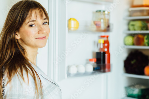 Young beautiful brunette woman in pajamas searching for food in the fridge and looking at camera.
