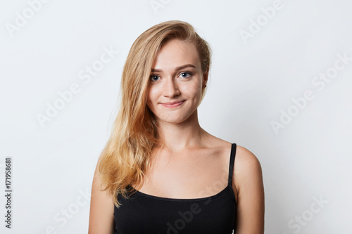 Adorable blue eyed female with long straight light hair, wears casual black t-shirt, looks happily into camera as going to walk with boyfriend, has good mood, isolated over white studio background