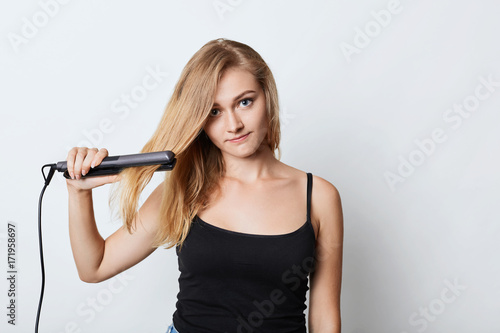 Image of blonde female in black clothing uses hair straightener, tries to do hairdo before going to party. Woman does hairstyle at home, uses hair iron. Female seller advertises beauty supplies