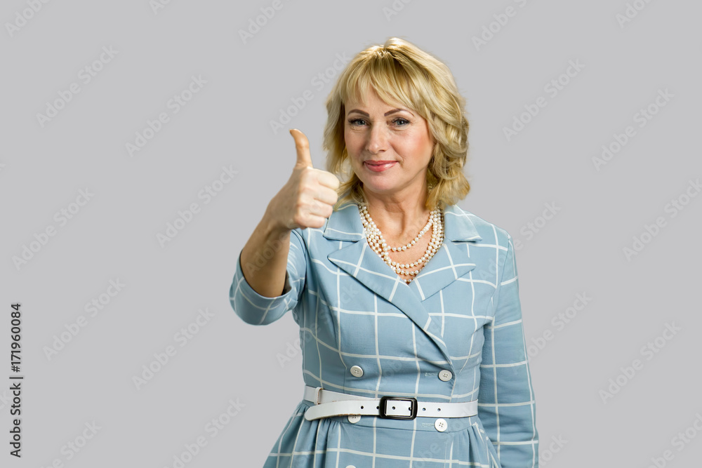Older elegant woman raised thumb up. Smiling white-skin lady raised her thumb up while standing on grey background close up. Older elegant woman raised thumb up. Smiling white-skin lady raised her thumb up while standing on grey background close up.