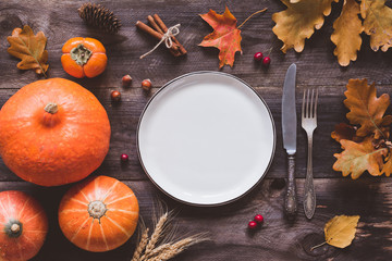 Autumn Halloween or Thanksgiving day table setting. Fallen leaves, pumpkins, spices, empty plate and vintage cutlery on wooden table. Top view, toned image