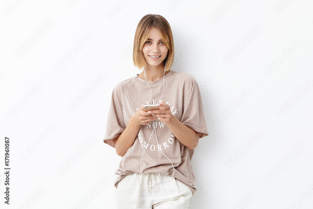 Waist up portrait of attractive young female adult wears oversized t-shirt, looks with delightful expression, going to run, listens music in earphones, isolated on white. People, lifestyle concept Waist up portrait of attractive young female adult wears oversized t-shirt, looks with delightful expression, going to run, listens music in earphones, isolated on white. People, lifestyle concept