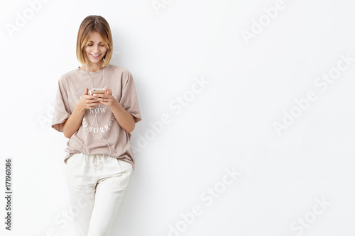 Teenge girl going to university, holds mobile phone, being glad to recieve message from boyfriend, listens to pleasant melody in earphones, isolated on white wall with copy space for your advertisment
