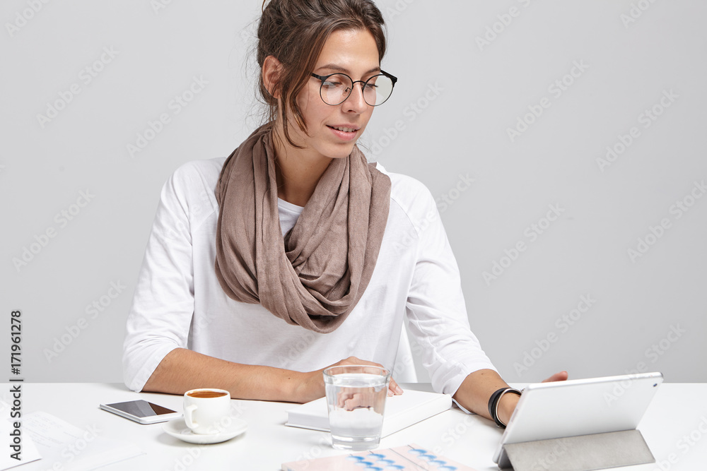 Stylish business lady wears glasses, sits at work place, uses modern digital tablet for preparing business report, drinks coffee during break, makes arrangment with business partners and companions Stylish business lady wears glasses, sits at work place, uses modern digital tablet for preparing business report, drinks coffee during break, makes arrangment with business partners and companions