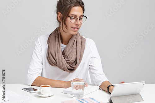 Stylish business lady wears glasses, sits at work place, uses modern digital tablet for preparing business report, drinks coffee during break, makes arrangment with business partners and companions