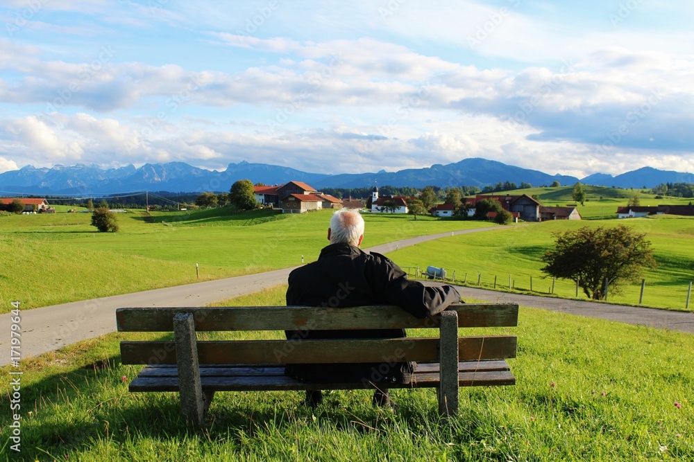 Älterer Mann sitzt auf einer Bank und genießt den Panoramablick auf die Berge, Allgäu, Bayern Älterer Mann sitzt auf einer Bank und genießt den Panoramablick auf die Berge, Allgäu, Bayern