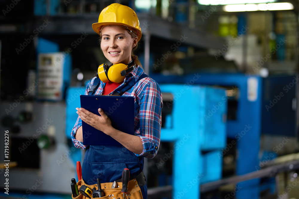 Waist-up portrait of attractive young technician in checked shirt and overall looking at camera with wide smile while carrying out inspection at production department of plant, blurred background Waist-up portrait of attractive young technician in checked shirt and overall looking at camera with wide smile while carrying out inspection at production department of plant, blurred background