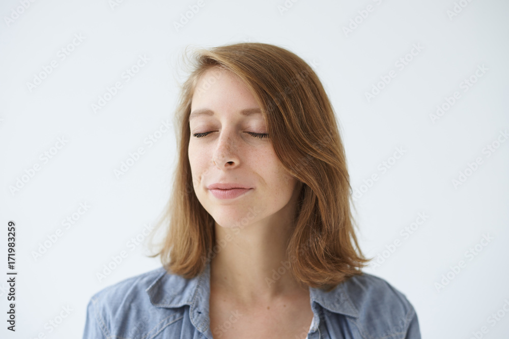 Headshot of positive young female posing at white studio wall with closed eyes while meditating, relaxing her mind, having calm and peaceful expression on her freckled face. Meditation and relaxation Headshot of positive young female posing at white studio wall with closed eyes while meditating, relaxing her mind, having calm and peaceful expression on her freckled face. Meditation and relaxation