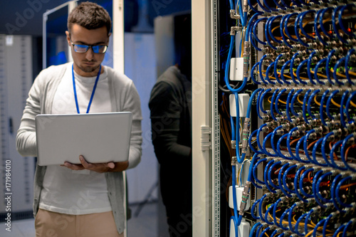 Portrait of young man with laptop standing by server cabinet while working with supercomputer in blue light photo