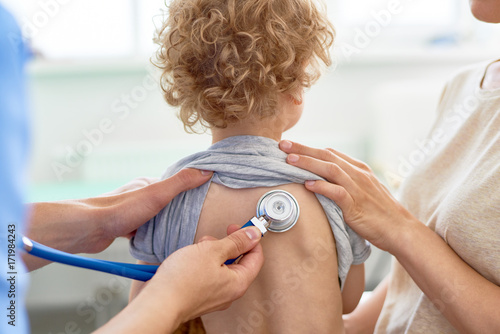 Unrecognizable male pediatrician examining curly little patient with help of stethoscope, blurred background photo