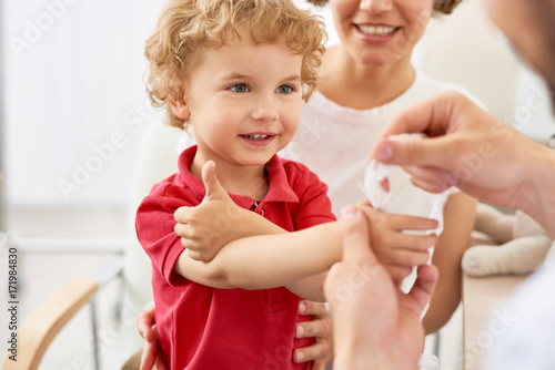 Fototapeta Waist-up portrait of smiling little boy showing thumb up while pediatrician putt