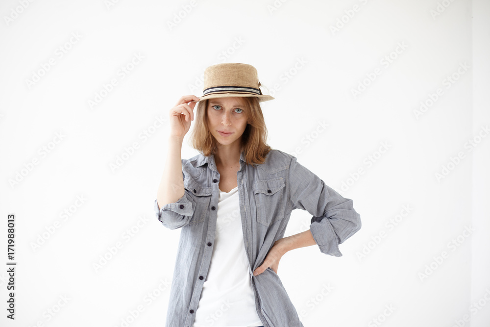 Style and fashion concept. Isolated studio shot of cool fashionable young European female model with blue eyes wearing vintage round hat and grey shirt over white t-shirt posing at blank wall