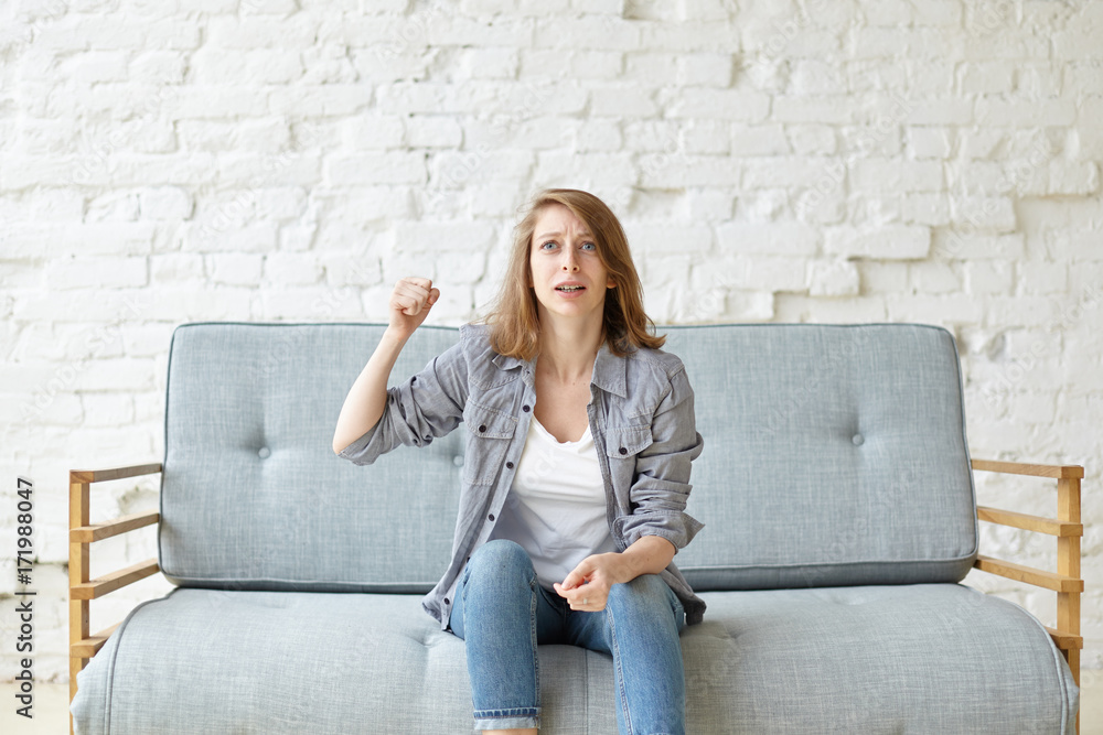 Indoor portrait of emotional excited young woman weaing casual clothes sitting on grey sofa at home alone and gesturing, cheering for local team while watching football match on TV in living room Indoor portrait of emotional excited young woman weaing casual clothes sitting on grey sofa at home alone and gesturing, cheering for local team while watching football match on TV in living room