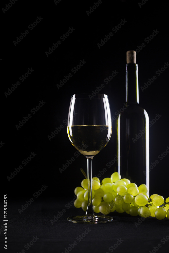 Glass of white wine in goblet on black background with dim light reflections. Copy space for text, wine tasting, bunch of fresh ripe yellow green grapes wines on dinner table, italian french Provence