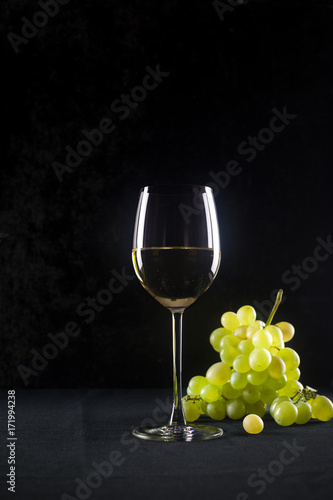 Glass of white wine in goblet on black background with dim light reflections. Copy space tasting, bunch of ripe yellow green grapes wines with bottle of italian french provence wine still life dinner