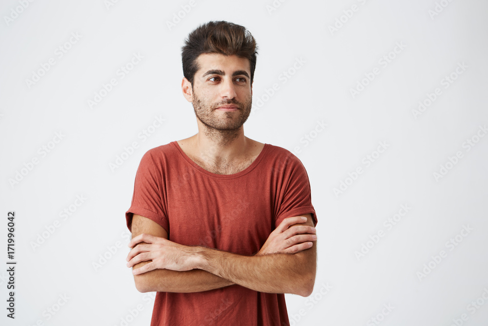 Young spanish hipster wearing red tshirt slightly smiling , keeping hands crossed, and meditative looking aside during magazine photoshoot. Beauty, people, youth and lifestyle concept Young spanish hipster wearing red tshirt slightly smiling , keeping hands crossed, and meditative looking aside during magazine photoshoot. Beauty, people, youth and lifestyle concept