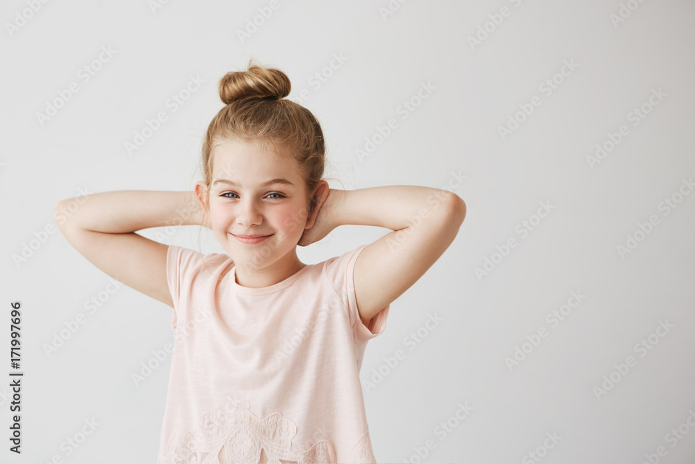Beautiful young miss with blue eyes and light hair in pink t-shirt posing with cute bun hairstyle and hands behind head for family photo. Beautiful young miss with blue eyes and light hair in pink t-shirt posing with cute bun hairstyle and hands behind head for family photo.