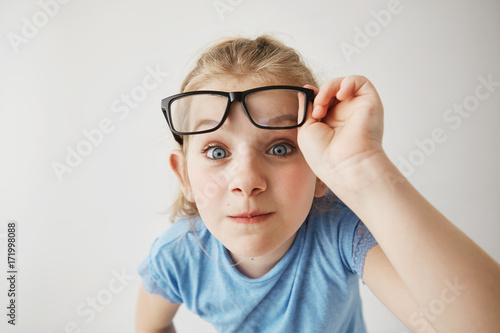 Close up portrait of cheerful small girl with blonde hair and blue eyes funny imitates adult person with glasses with surprised expression.