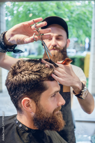 Dedicated male hairstylist using scissors and plastic comb while giving a cool haircut to a redhead bearded young man in a trendy beauty salon