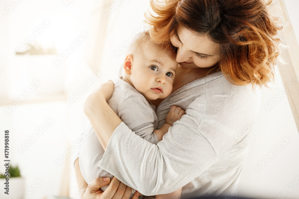 Cute newborn kid frightenedly looks aside while good-looking young mother gently huggs baby expressing her love and support. Family concept. Cute newborn kid frightenedly looks aside while good-looking young mother gently huggs baby expressing her love and support. Family concept.