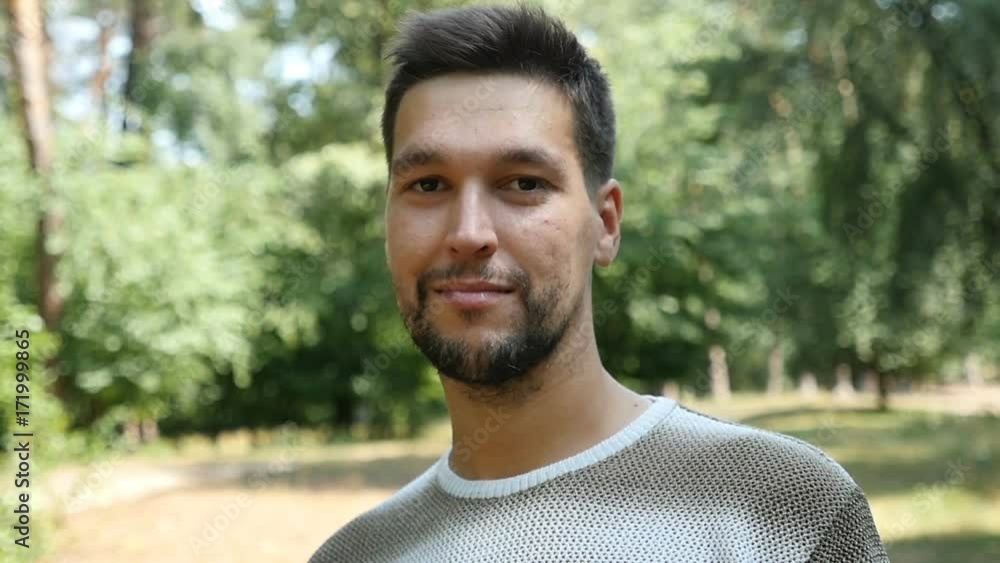 A closeup of a young bearded man who stands in a mixed forest and smiles sincerely in a relaxed way. He stands on a lawn full of sunny rays. A closeup of a young bearded man who stands in a mixed forest and smiles sincerely in a relaxed way. He stands on a lawn full of sunny rays.