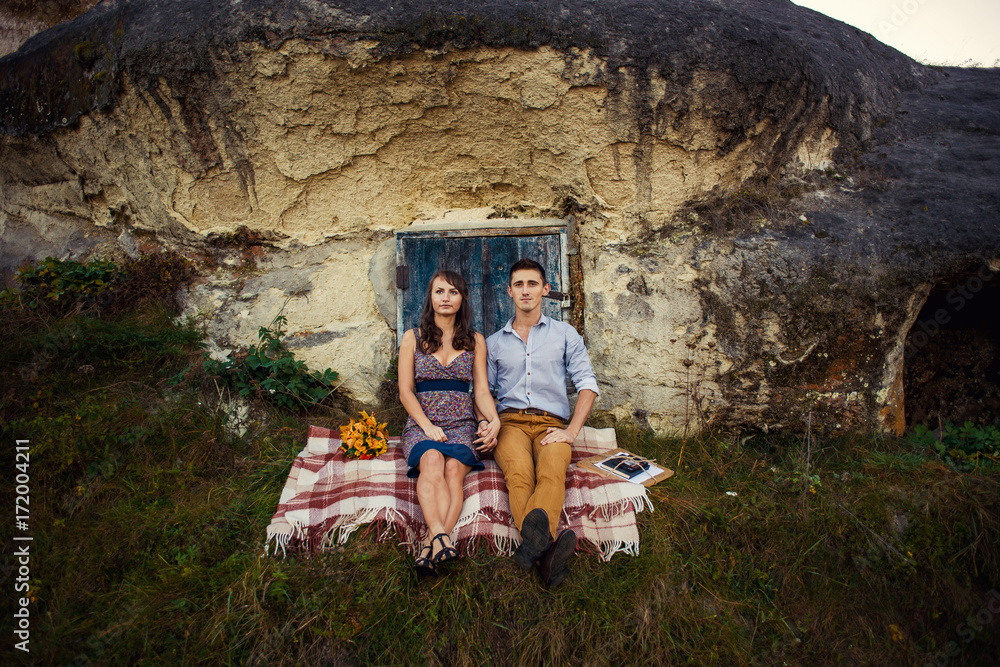 Sensual lovely couple sitting on the plaid near the rock with caves and blue wooden door.
