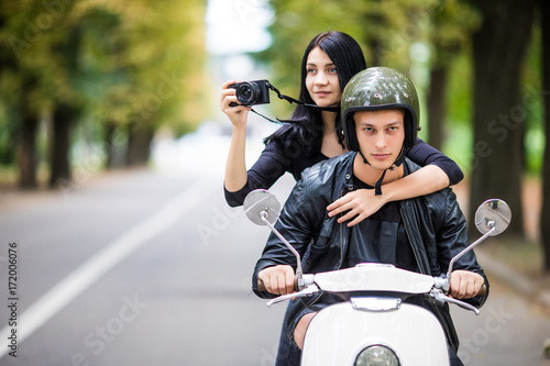 Happy tourist young couple on scooter ride in new city. Woman making photo on camera while riding outdoors