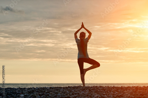 Girl at sunset practicing yoga at the seashore, back view