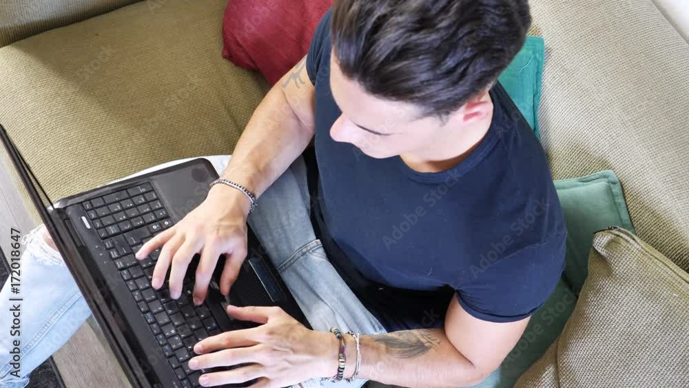 Attractive Young Man with Serious Expression, with Laptop on Couch, Working on his Start-up Business - Young Male College or University Student Doing Homework on Sofa Attractive Young Man with Serious Expression, with Laptop on Couch, Working on his Start-up Business - Young Male College or University Student Doing Homework on Sofa