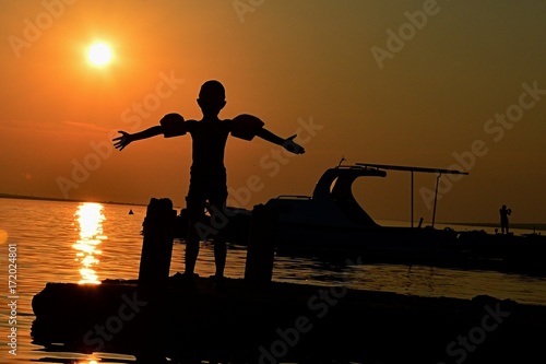 Silhouette of small boy spreading hands with swimming sleeves on arms towards sunset on molo in Croatia, Adriatic photo