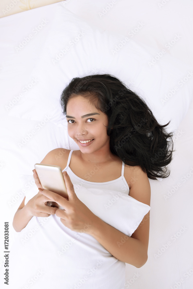 Smiling asian woman using mobile phone while hugging pillow Smiling asian woman using mobile phone while hugging pillow