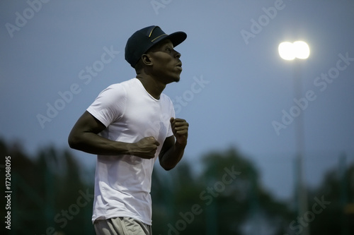 African male sportsman in a cap running on stadium track, he trains endurance