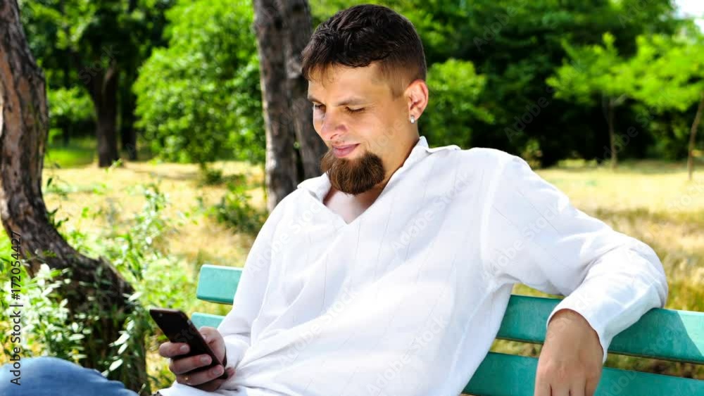 Man using business app on smart phone sitting on the bench in city park. Handsome young businessman communicating on smartphone smiling confident. Man using business app on smart phone sitting on the bench in city park. Handsome young businessman communicating on smartphone smiling confident.