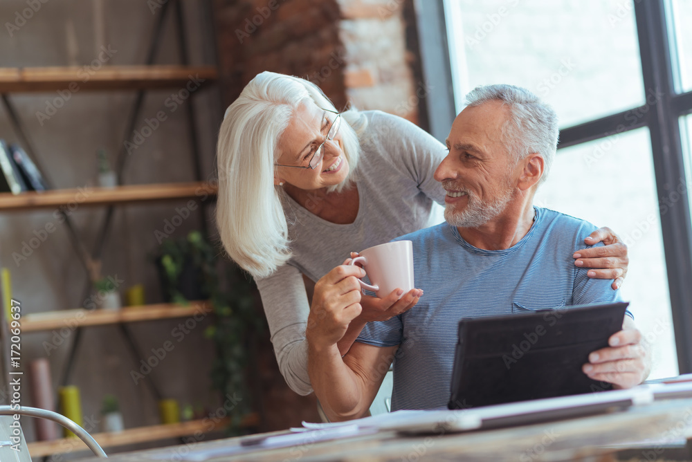 Positive aged wife giving a cup of tea to her husband - obrazy, fototapety, plakaty 