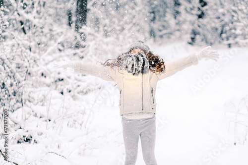 Happy little girl in white mittens and gray hat, standing in the snow, walking in winter park throws snow.