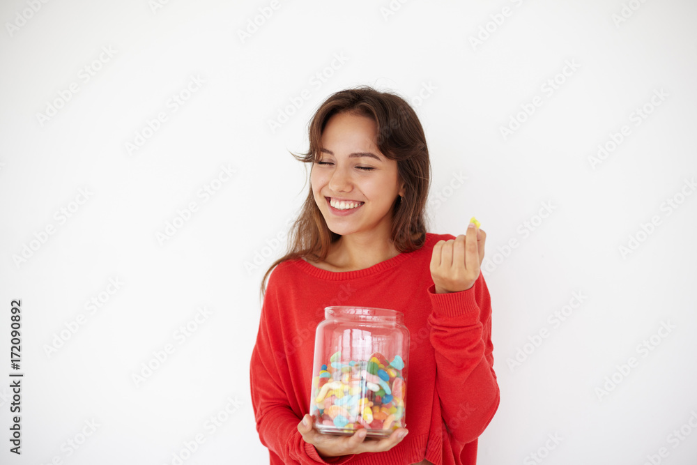 Joyful teenage girl wearing red sweater holding glass jar full of colorful jelly beans. Pretty woman enjoying unhealthy sugar food, closing her eyes in pleasure, posing against white wall background Joyful teenage girl wearing red sweater holding glass jar full of colorful jelly beans. Pretty woman enjoying unhealthy sugar food, closing her eyes in pleasure, posing against white wall background